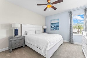 A bedroom with a white bed, a nightstand, a ceiling fan, a window with curtains, and a painting of the ocean.