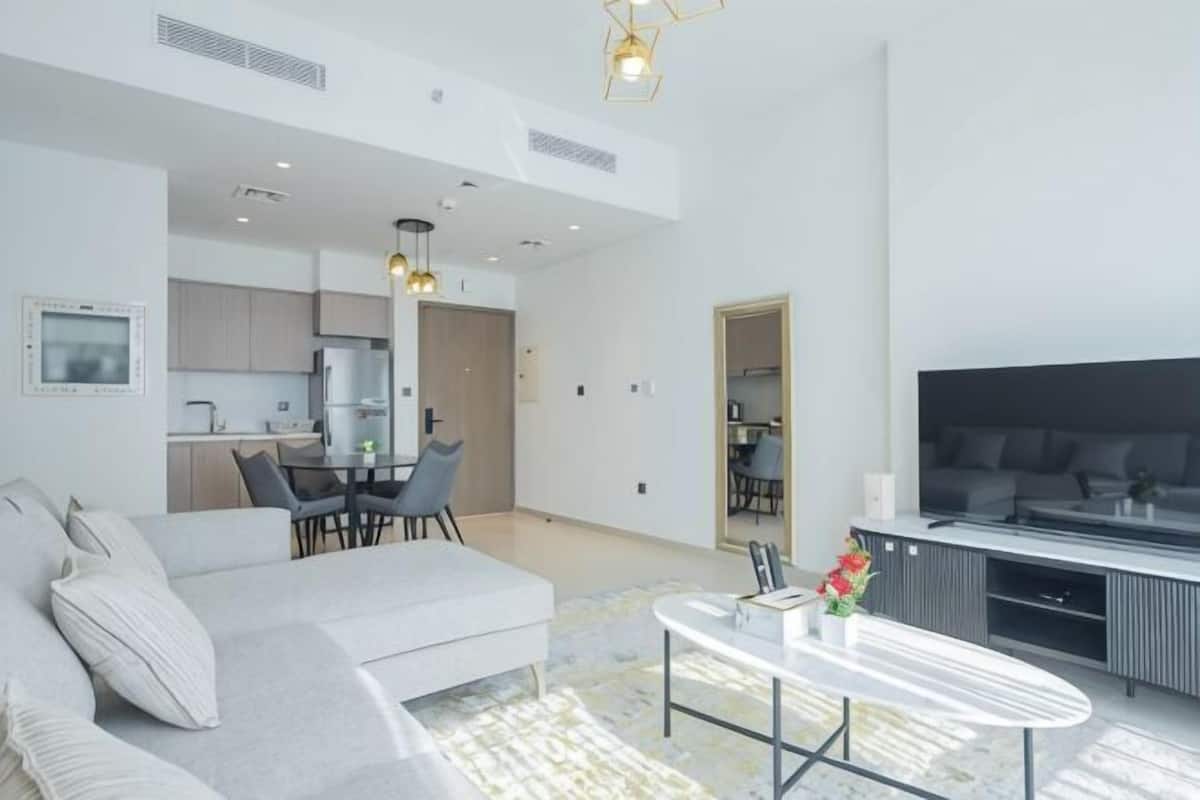 Un salón moderno con sofá, una mesa de centro y televisor. Cuenta con un comedor con sillas y una mesa. El ambiente tiene techos altos con iluminación empotrada.