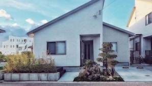 Una casa moderna de dos pisos con techo plano, un pequeño porche delantero y un jardín bien cuidado con un árbol bonsái.