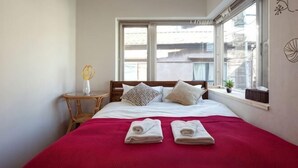 A bedroom with a bed, pillows, a red blanket, a window, and a small table with a vase. - Higashi Mukojima 4 Room (Tokyo)