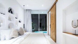 A bedroom with a bed, a wooden wardrobe, a white wall with shelves, and a sliding glass door leading to an outdoor area. - ULU 5 ·2br Villa (Bali)