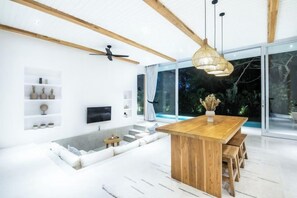 A modern interior with a wooden dining table, white sofas, a flat-screen TV, and a large window with a view of greenery. - ULU 5 ·2br Villa (Bali)