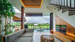 A modern living room with a sofa, a round wicker ottoman, and a TV on a wooden stand. - Kairos Villa Ubud (Bali)