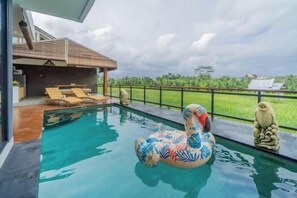 A swimming pool with a decorative flamingo float, a wooden deck with lounge chairs, and a view of a green landscape. - Kairos Villa Ubud (Bali)