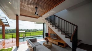 A modern living room with a large TV, wooden flooring, a staircase, and a view of a green landscape. - Kairos Villa Ubud (Bali)