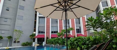 A swimming pool with a lounge area, including chairs and an umbrella, surrounded by greenery and a building with red and white patterned windows.