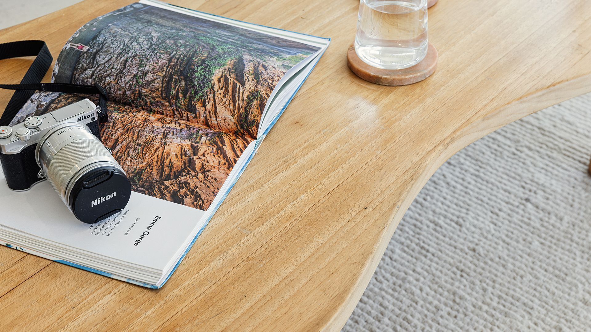 A Nikon camera, a magazine with a desert landscape, and a glass of water on a wooden table.