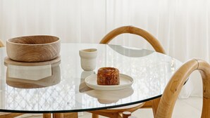 Una mesa redonda de vidrio con un cuenco de madera y un pastel en un plato, rodeada de sillas de madera.
