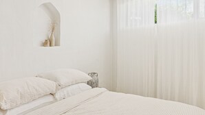 Un dormitorio minimalista con cama, almohadas, un jarrón y cortinas de gasa.
