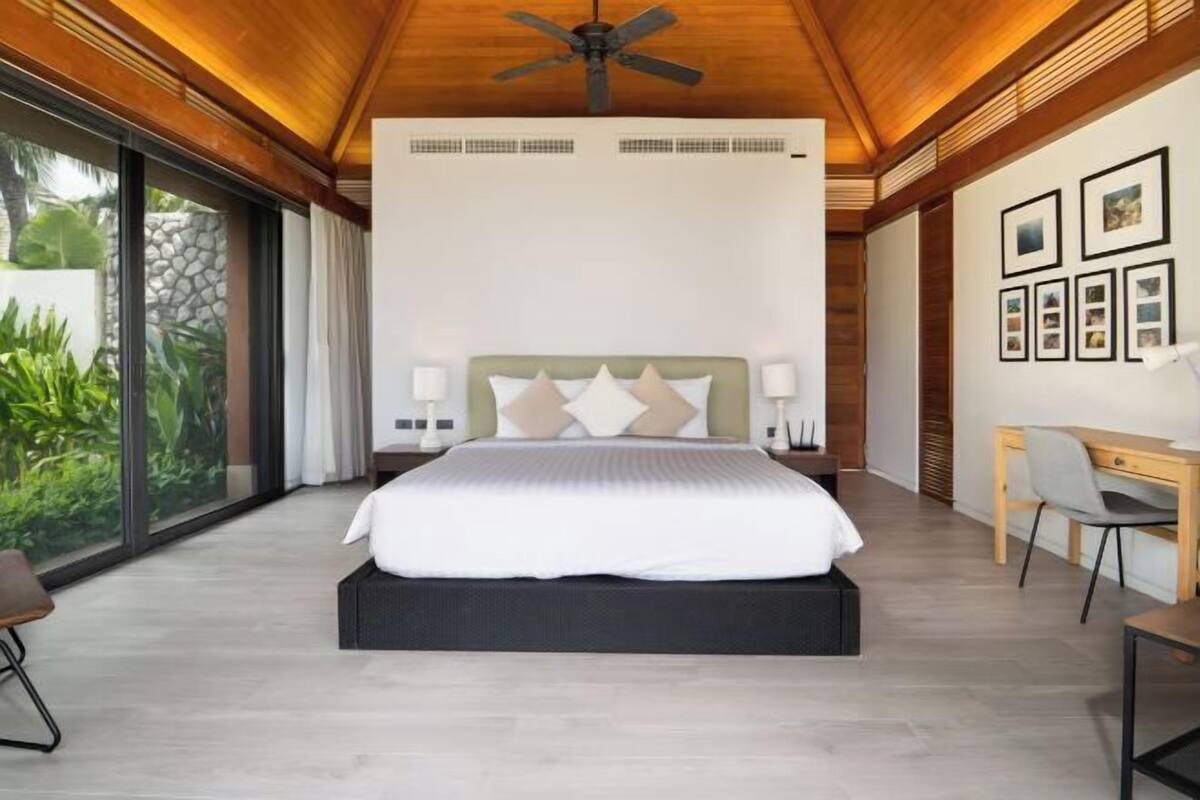 A modern bedroom with a large bed, a desk, and a chair, and a view of greenery outside.