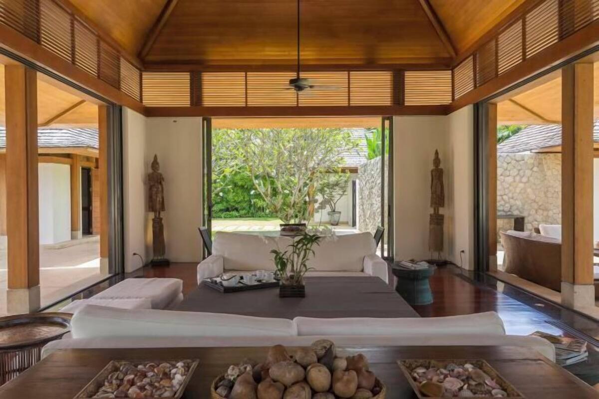 A spacious room with a large sofa, a wooden table with decorative items, and a view of a garden through large windows.