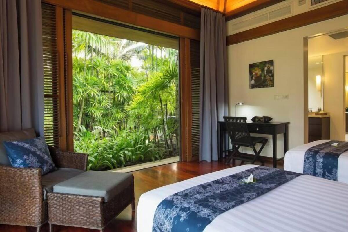 A hotel room with a large sliding glass door leading to a lush tropical garden, a bed with a patterned bedspread, a desk with a chair, and a small seating area with a cushioned bench.