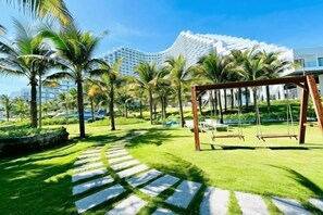 A pathway leading to a swing set in a well-maintained garden area with palm trees and a modern building in the background. - Resort CAM RANH DELUXE SEAVIEW (Cam Ranh)