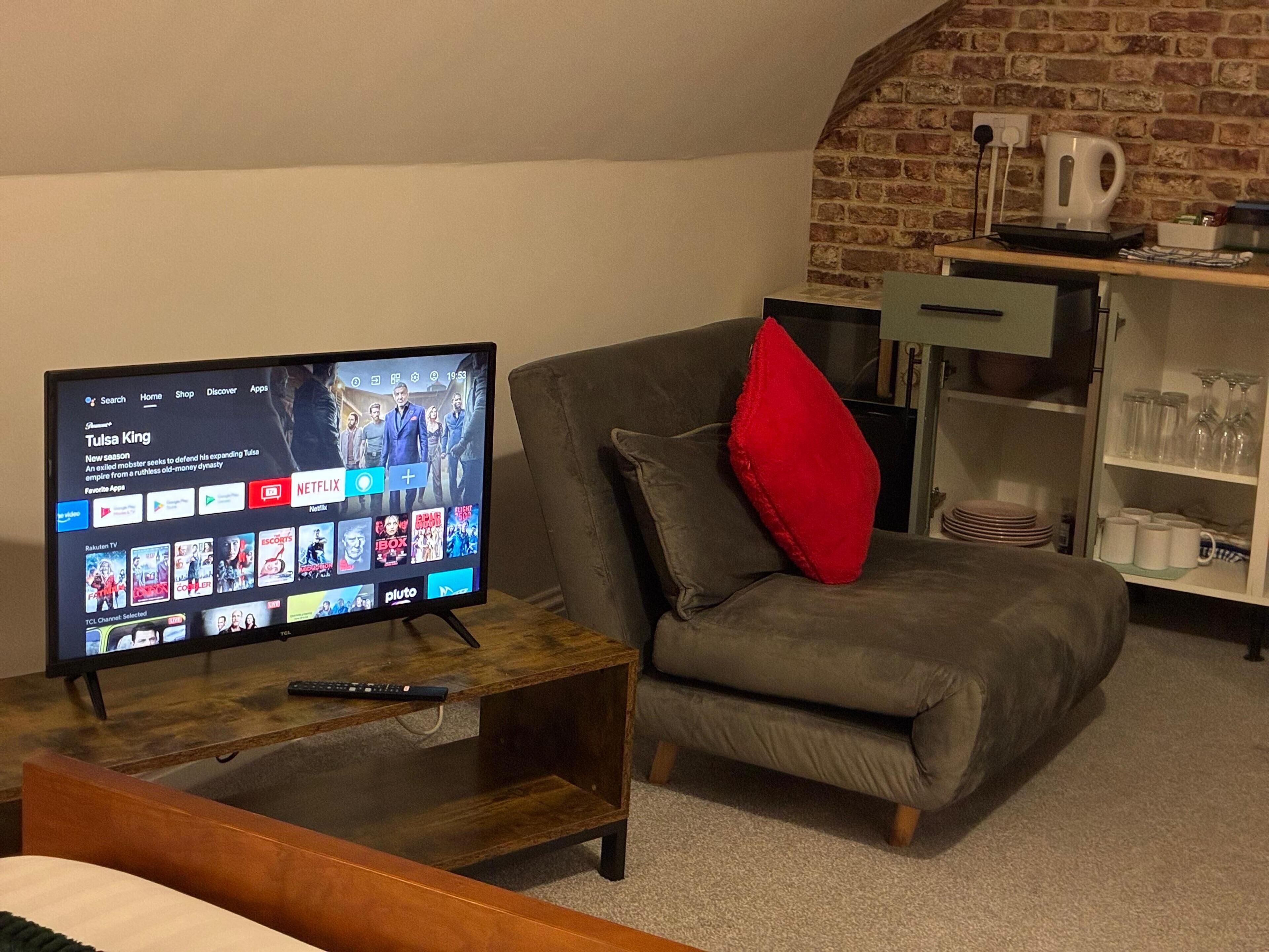 A living room with a flat-screen TV on a wooden stand, a grey armchair with a red cushion, and a shelf with glassware.