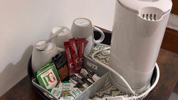 Eine Kaffeemaschine, eine Tasse, ein Löffel, eine Kaffeepackung, eine Teepackung, eine Zuckerpackung, eine Milchpackung und eine Kaffeeweißerpackung auf einem Tablett.