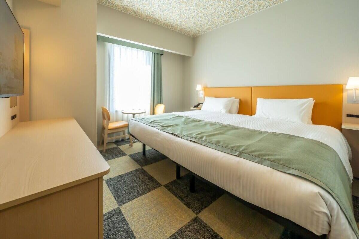 Una habitación de hotel con cama, escritorio, silla y ventana con cortinas.