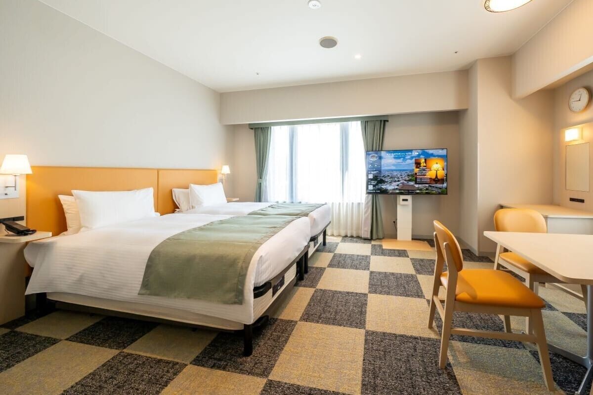 Habitación de hotel con cama, mesita de noche, lámpara, televisor y una pequeña zona de comedor con una silla y mesa.