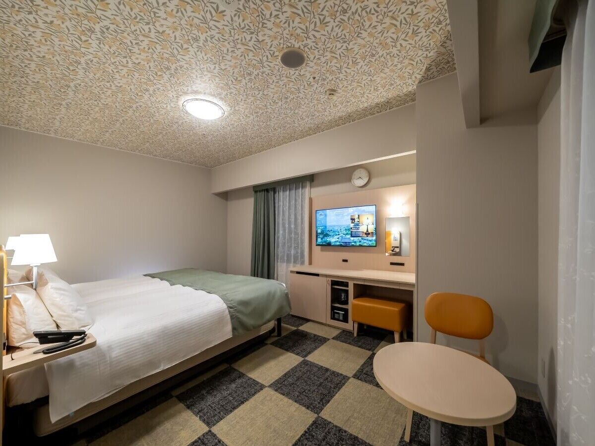 A hotel room with a bed, a desk with a computer, a chair, a small table, and a television.