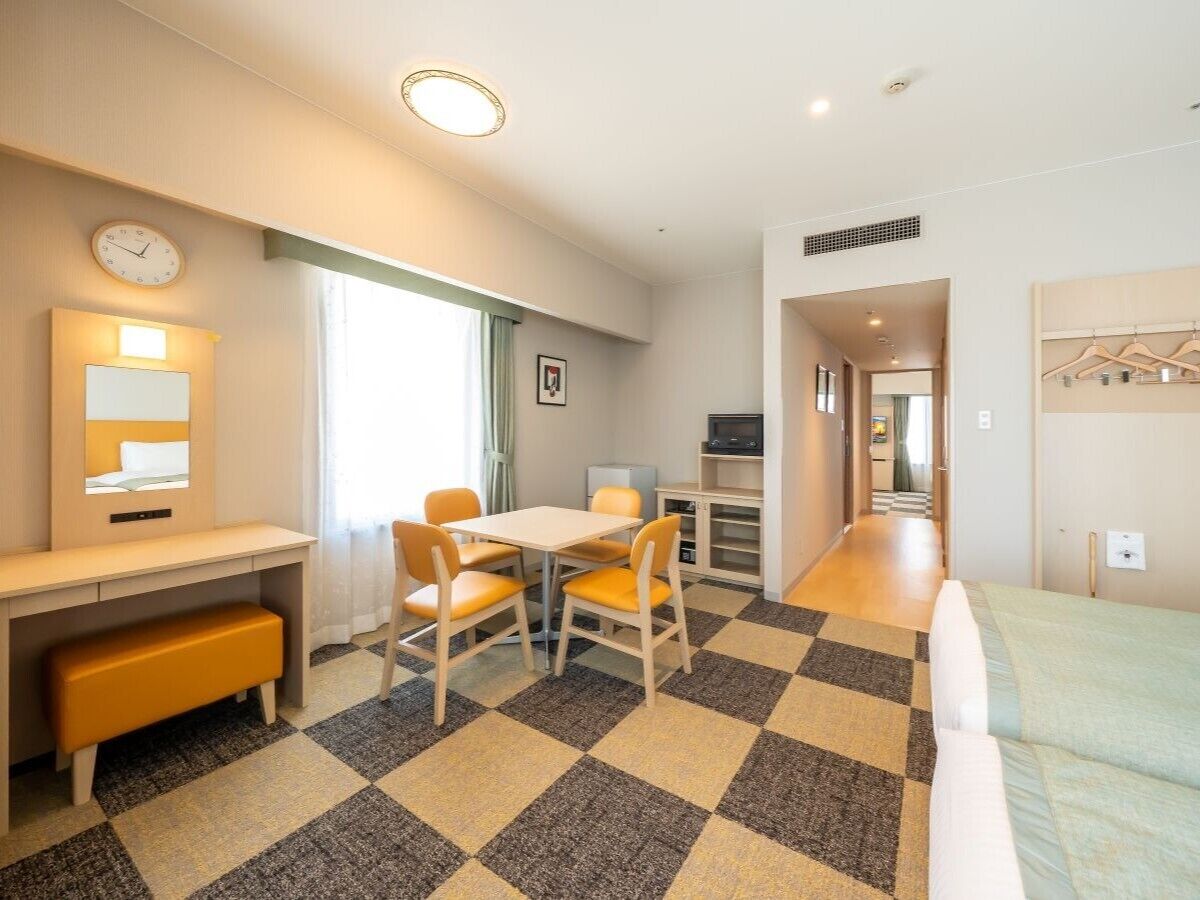 Ein Hotelzimmer mit einem Bett, einem Schreibtisch mit Stuhl, einem kleinen Tisch mit Stühlen, einer Uhr, einem Bild an der Wand und einem Schachbrettfußboden.