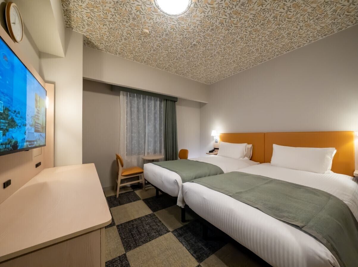 A hotel room with two beds, a desk, a chair, a TV, and a patterned ceiling.
