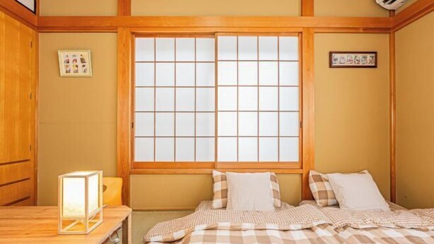 Una habitación tradicional japonesa con suelo de tatami, una cama baja de madera con ropa de cama a cuadros, una ventana con pantalla shoji y una pequeña mesa con una lámpara.