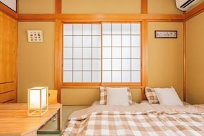 A traditional Japanese-style room with tatami flooring, a low wooden bed with a checkered bedding, a shoji screen window, and a small table with a light fixture.