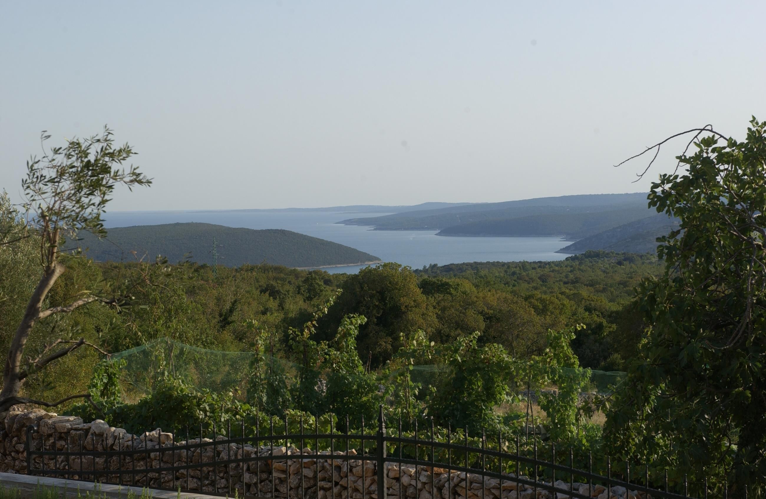 Una vista panorámica de un paisaje costero con un muro de piedra y una cerca de metal en primer plano, que conducen a una masa de agua y colinas lejanas.