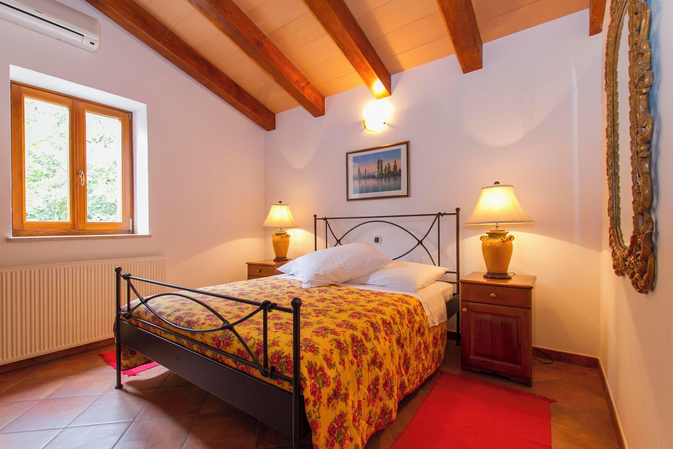 A bedroom with a bed, two bedside tables with lamps, a window, a radiator, and a painting on the wall.