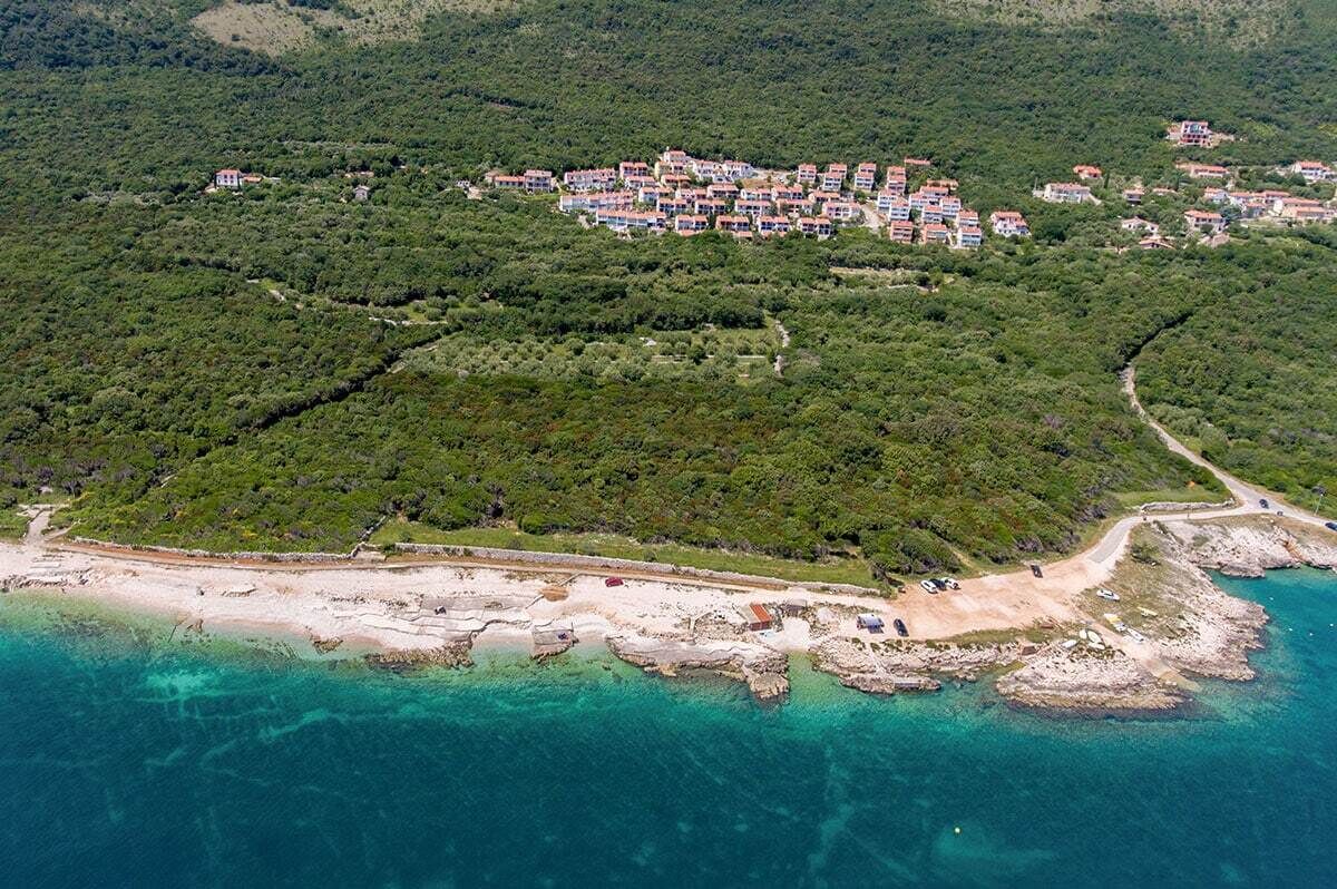 An aerial view of a coastal area with a cluster of buildings surrounded by dense greenery and a sandy beach with clear turquoise waters.