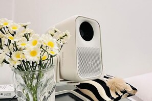 A white portable speaker with a perforated front panel, placed next to a glass vase with white flowers. - Five House Silom 2 Townhouse (Bangkok)