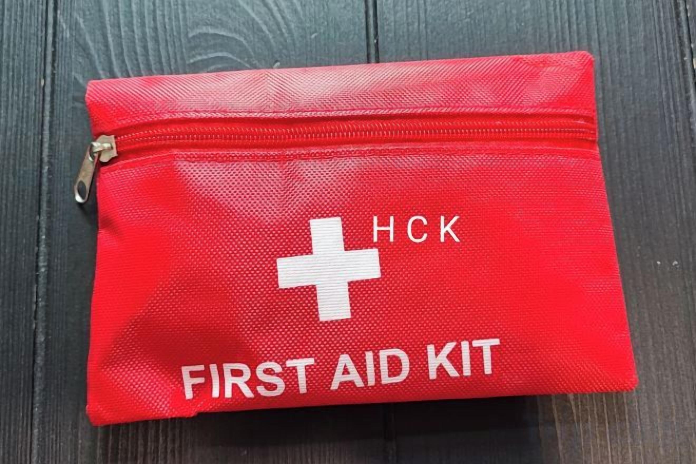 Une trousse de premiers secours rouge, ornée d’une croix blanche et portant l’inscription « HCK FIRST AID KIT », est posée sur une surface en bois sombre.