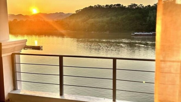 A balcony with a view of a sunset over a lake, a boat, and a forested hill. - Sokview Residence (Luang Prabang)