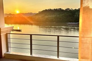 A balcony with a view of a sunset over a lake, a boat, and a forested hill. - Sokview Residence (Luang Prabang)