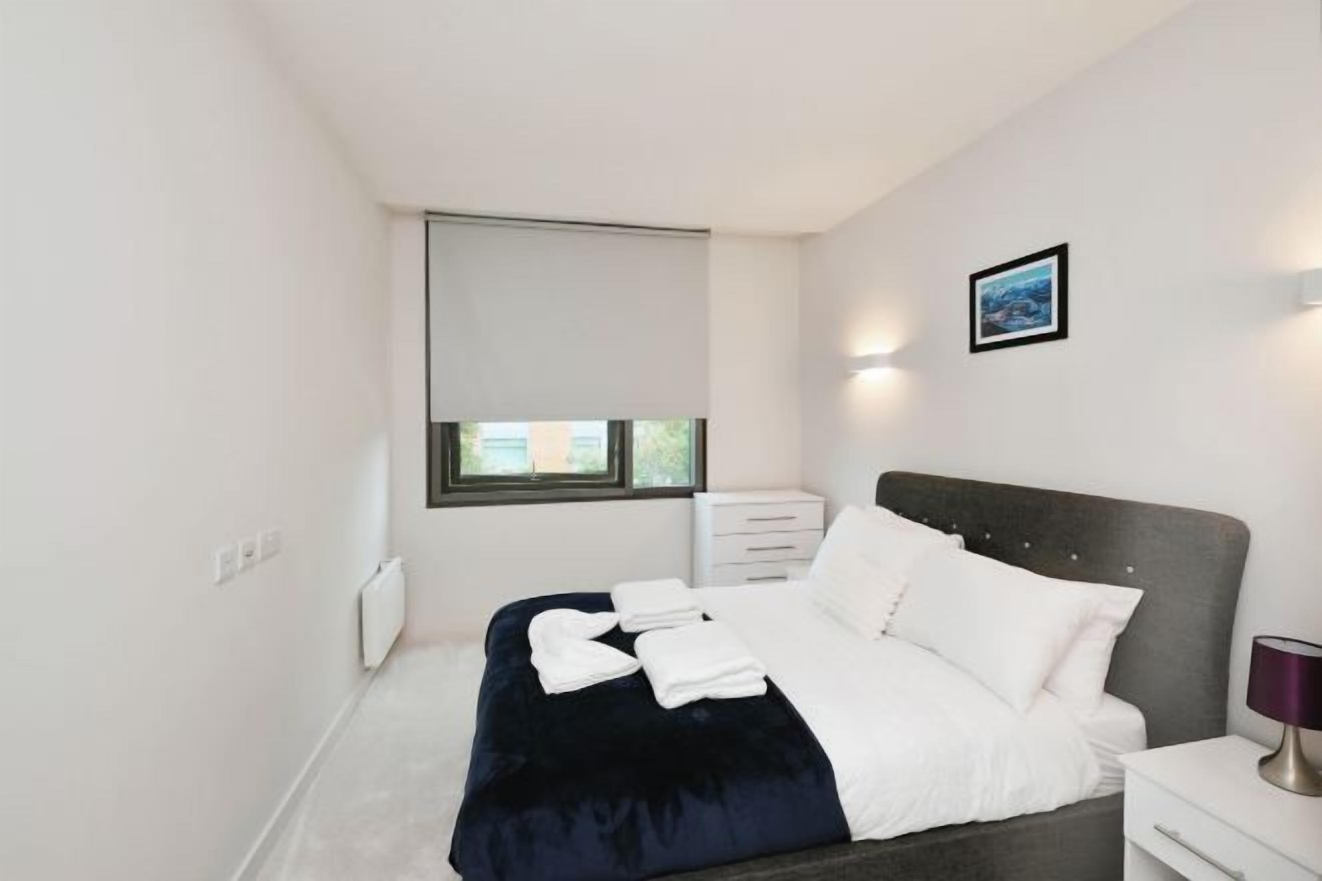 A modern bedroom with a dark blue bedspread, white pillows, and a bedside table with a lamp.