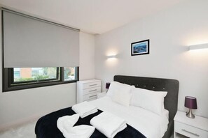 A modern hotel room with a bed, bedside table, and a window with blinds. - Modern 1 Bed Apt Near Ascot- with parking (Bracknell)