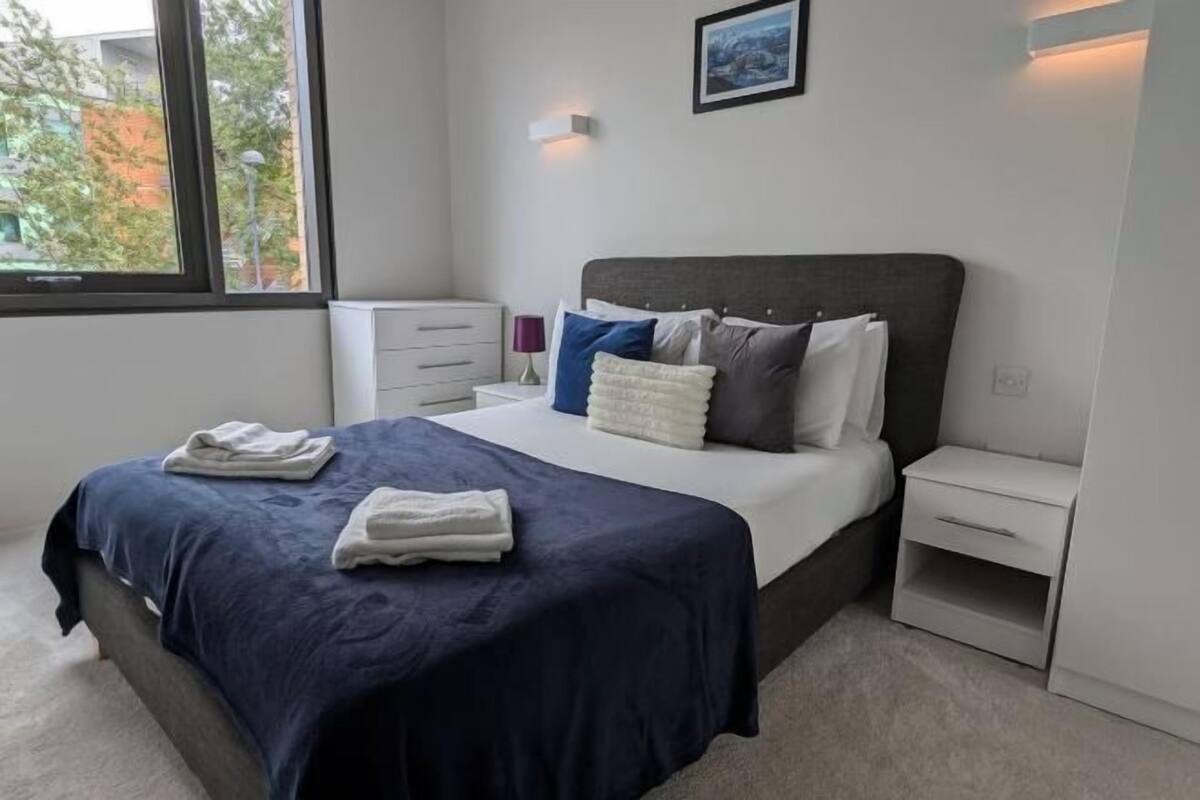Una cama bien tendida con una colcha azul oscuro y toallas blancas, una mesita de noche con una lámpara, y un pequeño mueble auxiliar con un cajón.