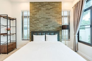 A bedroom with a stone wall, a bed with white bedding, a nightstand, a lamp, and a window with curtains. - Me&You Pool Villa Hua Hin (Cha-am)