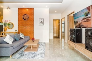 A modern living room with a gray sofa, a coffee table, a brick accent wall, a large TV, and a pair of speakers.