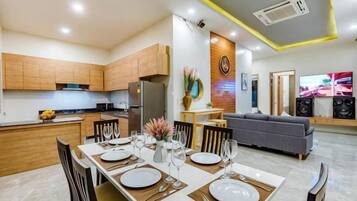 A modern kitchen with wooden cabinets, a dining area with a table set for four, and a living area with a sofa and TV.