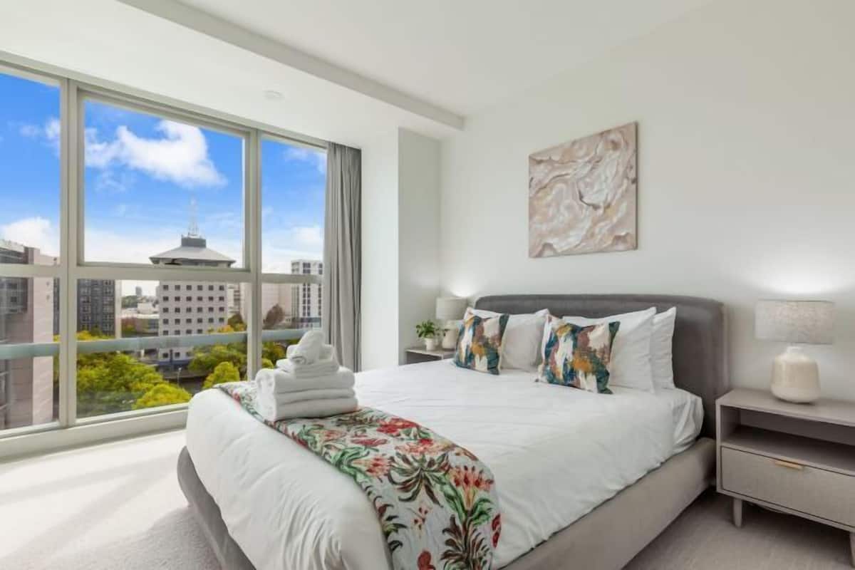A modern hotel room with a large window, a bed with white bedding, a gray headboard, and a nightstand with a lamp.