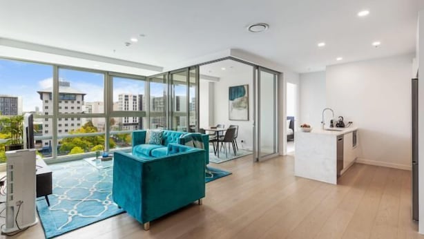 Een modern appartement met een groot raam, een blauwgroene bank, een keukenhoek en een eethoek.