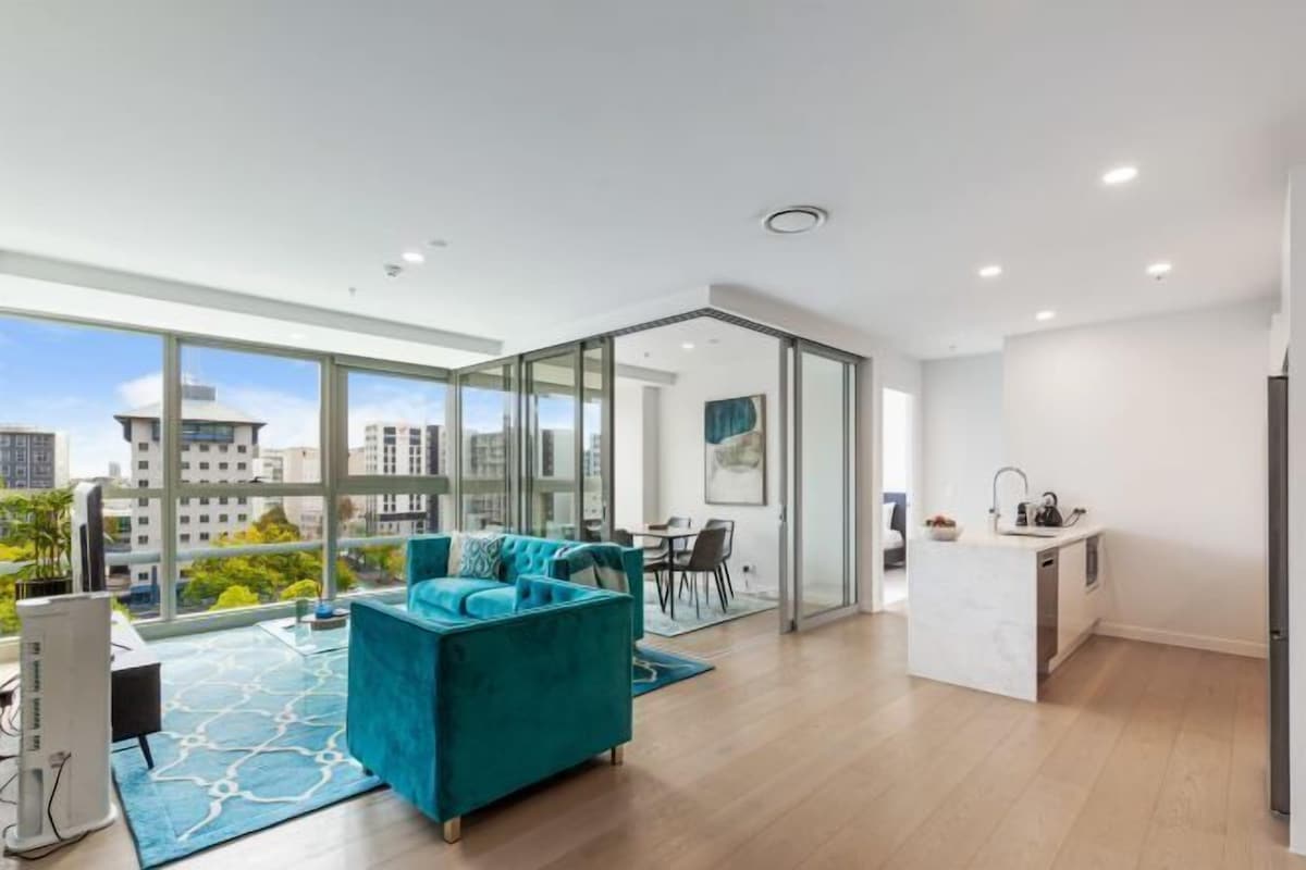 A modern apartment with a large window, a teal sofa, a kitchen area, and a dining space.