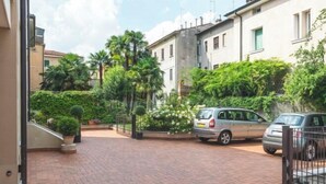 A paved driveway leading to a gated entrance with parked cars, surrounded by lush greenery and residential buildings. - Juliet House (Verona)