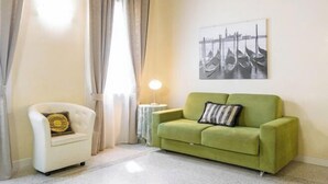 A room with a green sofa, a white armchair, a lamp, and a painting of gondolas. - Juliet House (Verona)