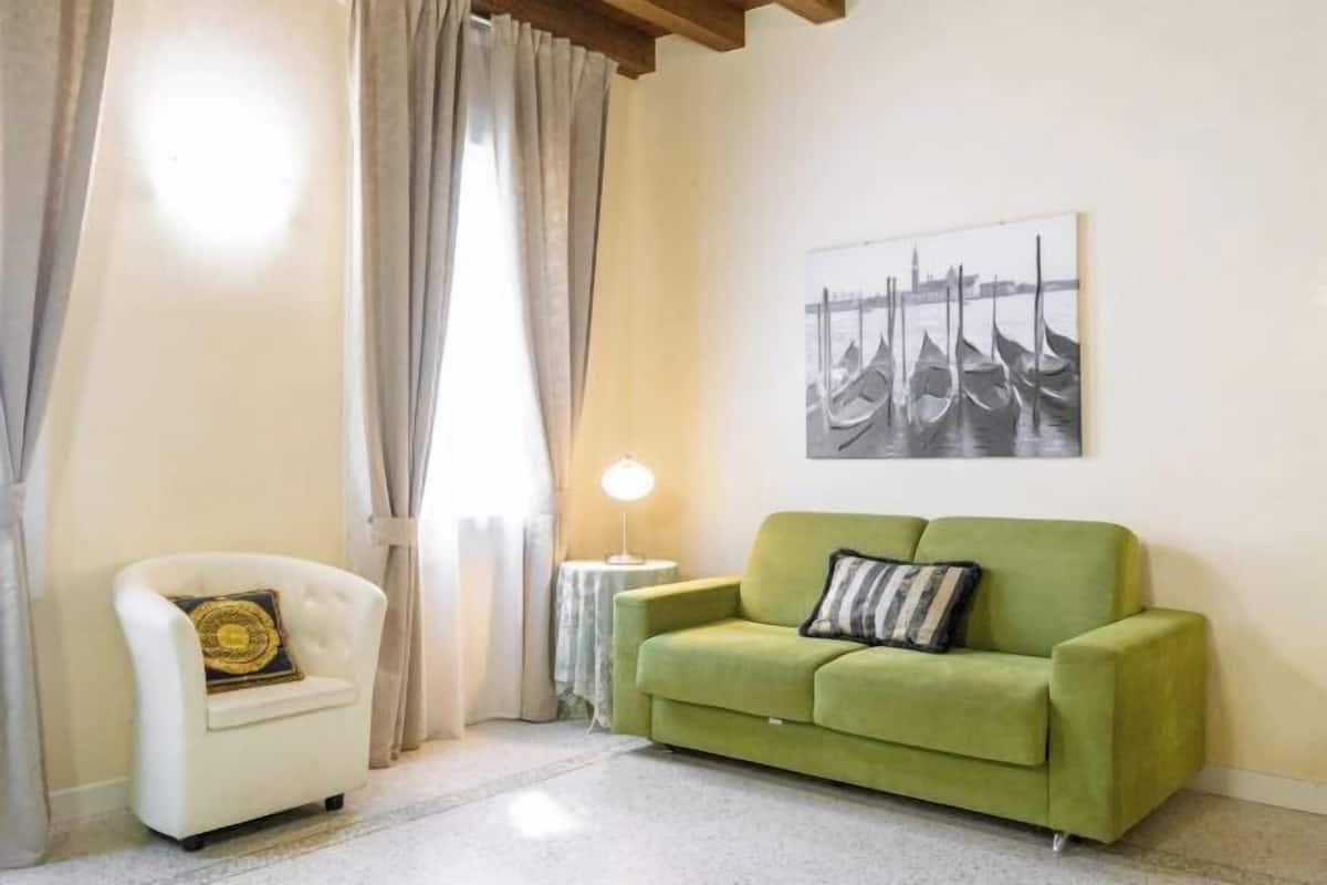 A room with a green sofa, a white armchair, a lamp, and a painting of gondolas.