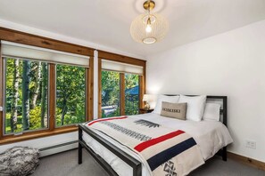A bedroom with a bed, a nightstand, a lamp, and a window with a view of trees. - Evergreen Escape – Home for 10 (Vail)