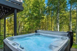 A hot tub with a metal railing and a wooden deck, surrounded by a forest with tall trees.