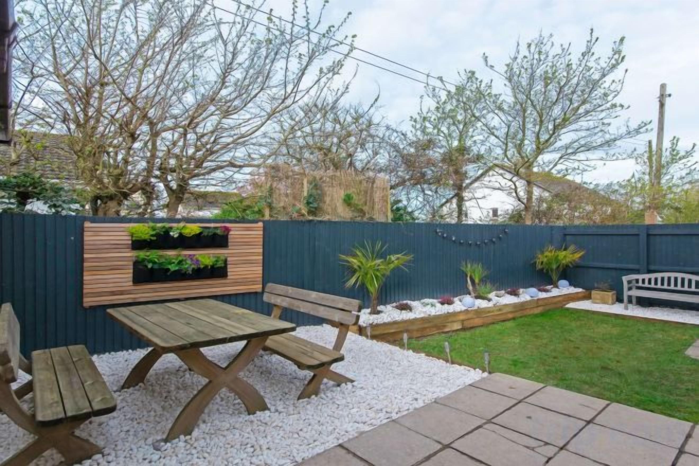 Un patio con una mesa y bancos de madera, un cantero con plantas y una cerca azul.