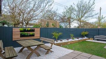 Une terrasse avec une table et des bancs en bois, un parterre fleuri et une clôture bleue.