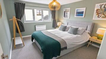 Une chambre à coucher avec un lit, des tables de chevet, une fenêtre avec des rideaux, un radiateur et des œuvres d’art accrochées aux murs.